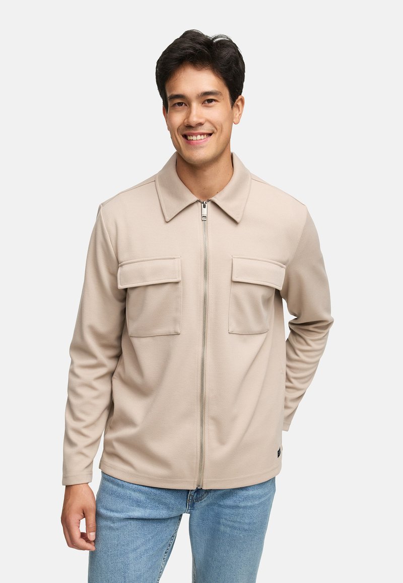 Beige Zip-Jacke mit Reißverschluss und Kragen, strukturiertem Stoff, zwei Brusttaschen und langen Ärmeln, kombiniert mit hellblauen Jeans.