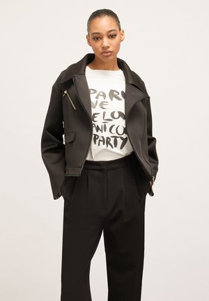 Schwarze Jacke mit strukturiertem Kragen und Frontreißverschluss, getragen über einem weißen Grafik-T-Shirt. Schwarze Hose mit Falten vervollständigt den Look.