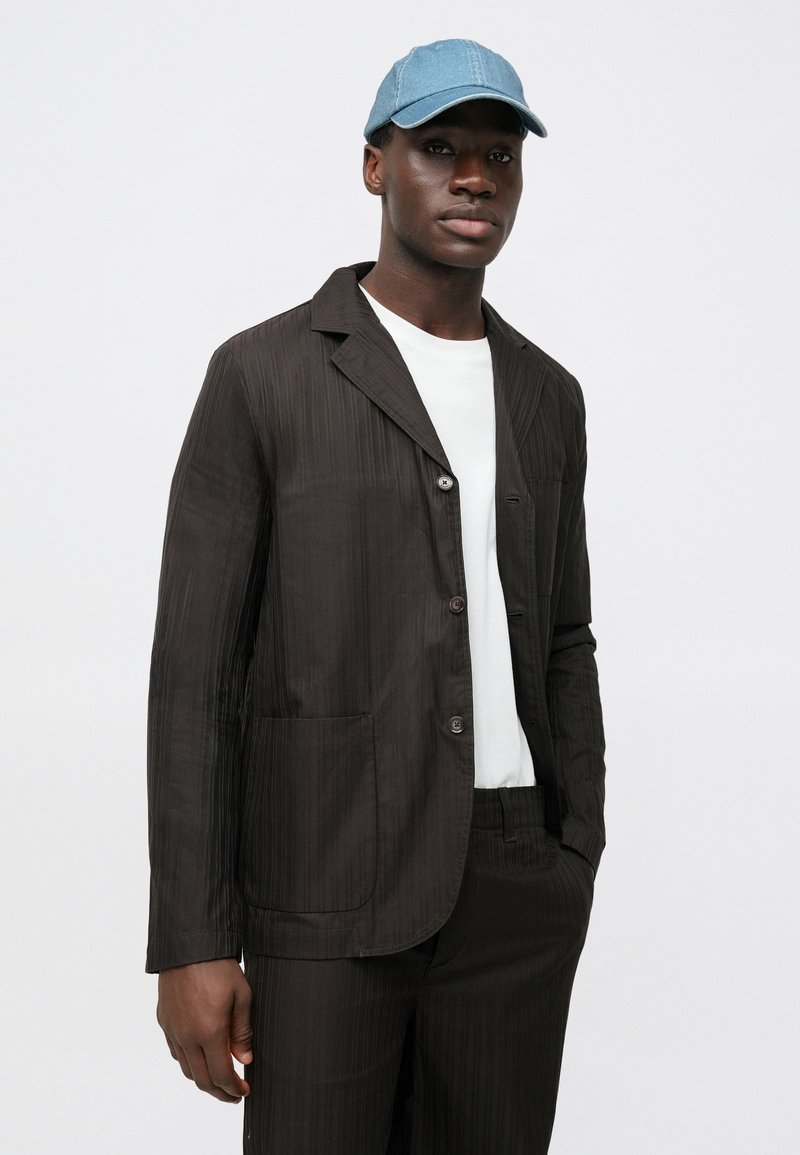 Bruine blazer met krijtlijnen, voorzien van een reverskraag, knoopsluiting en zijzakken, gedragen over een wit overhemd, gecombineerd met een lichtblauwe pet.
