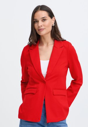 Femme aux cheveux foncés mi-longs, portant des boucles d’oreilles anneaux, un blazer rouge vif, un haut blanc et un jean bleu sur un fond clair uni.