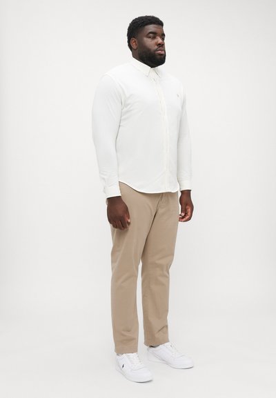 Chemise blanche à manches longues avec un col, pantalon beige et sneakers blancs, se tenant devant un fond uni. Design simple et couleurs neutres.