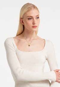 Gerippte Langarmoberteil in Creme mit quadratischem Ausschnitt. Eine goldene Anhängerhalskette liegt am Schlüsselbein. Glatte Textur, eng anliegendes Design.