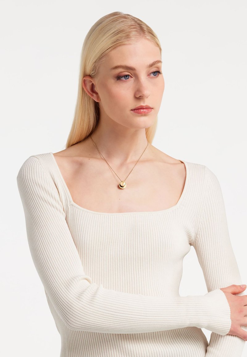 Gerippte Langarmoberteil in Creme mit quadratischem Ausschnitt. Eine goldene Anhängerhalskette liegt am Schlüsselbein. Glatte Textur, eng anliegendes Design.
