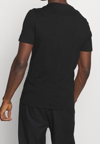 Muž oblečený v jednoduchém černém tričku s krátkým rukávem a černých kalhotách, zachycený ze zadu na jednobarevném světlém pozadí.