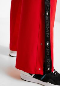 Pantalones deportivos rojos con botones a los lados y la marca Nike Pro en negro. Se llevan con zapatillas deportivas negras con suela blanca y detalles en azul.