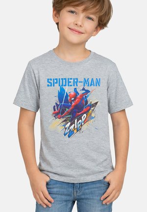 Jonge jongen draagt een grijs Spider-Man grafisch t-shirt en een blauwe spijkerbroek, lacht met handen in zakken tegen een effen witte achtergrond.