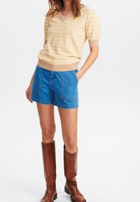 Kvinde iført gul og pink ternet kortærmet sweater, blå denimshorts med store forlommer og brune knælange læderstøvler.