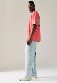 Kortærmet koralfarvet t-shirt med grafisk detaljer på ryggen, parret med lyseblå afslappede jeans og beige sneakers.