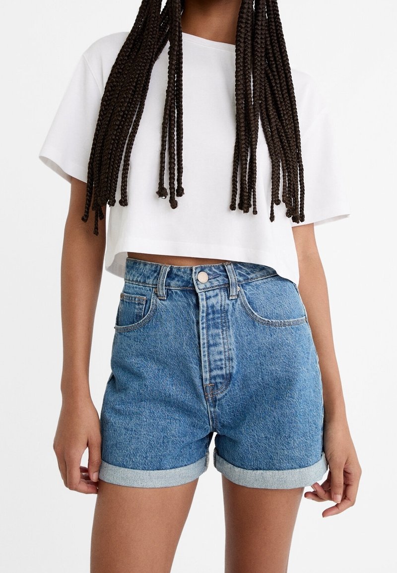 Personne portant un t-shirt court blanc et un short en jean bleu taille haute avec des revers, debout devant un fond blanc uni.