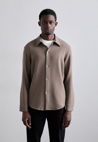 Chemise à manches longues beige avec un col plat, texture douce, ourlet arrondi, avec des boutons blancs, portée sur une chemise blanche.