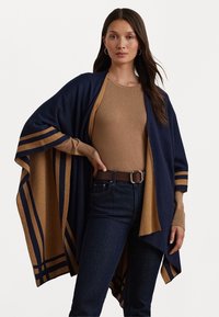 Cardigan aperto blu navy e beige con accenti a righe, realizzato in materiale morbido. Indossato su una maglietta beige, abbinato a jeans scuri e una cintura marrone.