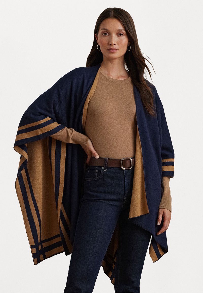 Cardigan aperto blu navy e beige con accenti a righe, realizzato in materiale morbido. Indossato su una maglietta beige, abbinato a jeans scuri e una cintura marrone.