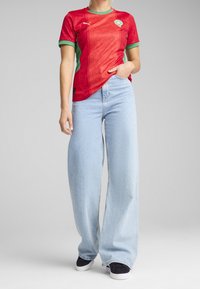 Osoba ubrana w czerwono-zielony strój piłkarski Maroka, jasnoniebieskie szerokie dżinsy i czarne sneakersy, stojąca z jedną ręką w kieszeni.