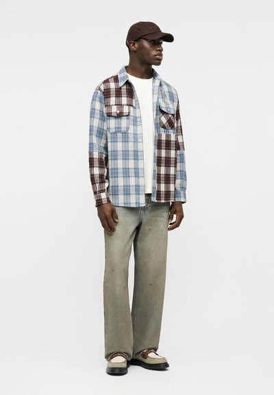 Muž v hnědém a modrém kostkovaném bundovém košili, bílém tričku, volných vybledlých šedých džínách, béžovo-hnědých botách a hnědé čepici.