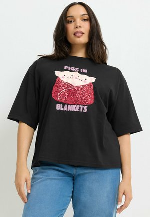 Femme portant un t-shirt noir oversize avec deux cochons enveloppés dans une couverture rouge pailletée et le texte "Pigs in Blankets" à l'avant.