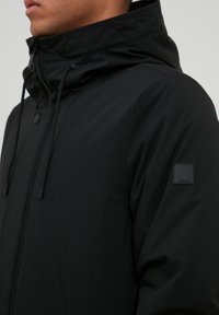 Chaqueta negra con capucha que presenta un cuello acolchado, botones a presión en la parte frontal, cordones ajustables y un pequeño parche de logo en la manga.