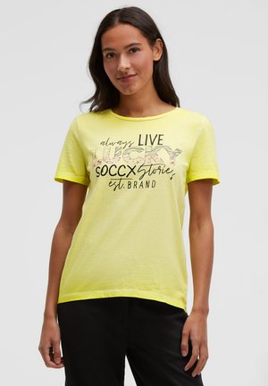 Woman with long dark hair wearing a yellow T-shirt with black pants, T-shirt reads "always LIVE LUCKY SOCCX Stories est. BRAND".