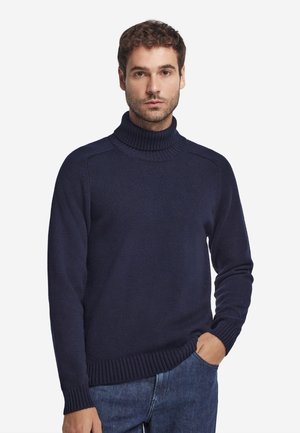 Man draagt een marineblauwe coltrui met geribbelde boorden en zoom, gecombineerd met een blauwe jeans, staand met één hand in de zak.