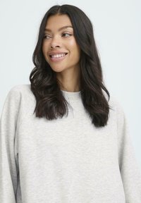 Sudadera gris clara con una textura suave y mangas raglán, que presenta un cuello redondo y un ajuste relajado, llevada por una persona con cabello oscuro.