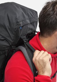 Schwarzer Rucksack mit texturiertem Finish, verstellbaren Riemen und einem oberen Deckel. Die Person trägt ein rotes Shirt und greift mit einer Hand nach einem Riemen.