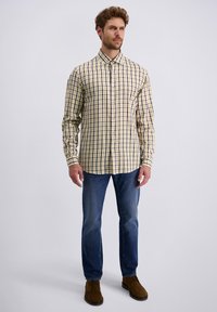 Mann steht mit einem gelben karierten Hemd, blauen Jeans und braunen Wildlederschuhen vor einem einfarbigen hellen Hintergrund.