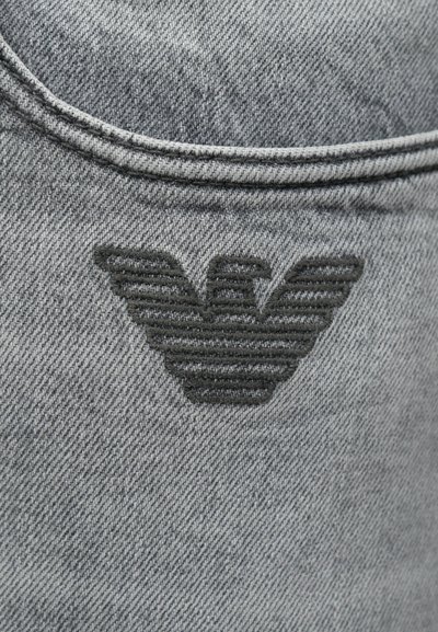Το γκρι τζιν ύφασμα διαθέτει ένα ραμμένο μαύρο λογότυπο που μοιάζει με στυλιζαρισμένο 'W' με οριζόντιες γραμμές, τοποθετημένο σε μία πίσω τσέπη.