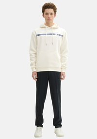 Sudadera blanca con cordones, franjas azules horizontales en el pecho, bolsillo frontal; combinada con pantalones oscuros y zapatillas blancas.