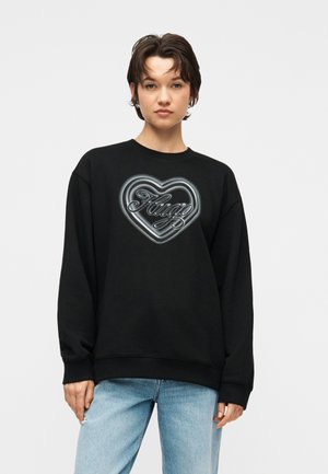 Junge Person mit kurzen dunklen Haaren, die einen schwarzen Pullover mit einem glänzenden Herzdesign und dem Schriftzug "Fuego" trägt, kombiniert mit hellblauen Jeans.