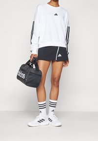 Vit långärmad sweatshirt med tre svarta ränder, svart kjol med dragkedja, svart Adidas duffelväska, vita skor med svarta detaljer, randiga strumpor.