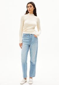 Creme ribbet langærmet top, høj hals; lys blå lige ben jeans; hvide vævede slip-on sko; neutral baggrund.