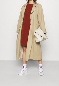 Vrouw in een beige trenchcoat over een roestkleurige jurk, met witte sneakers, houdt een grote geweven crèmekleurige clutch in haar hand, staand binnen.