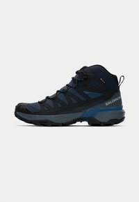 Mørkeblå og sorte middeishøye turstøvler med robust såle, snøring, trekkstroppe bak og "Salomon"-merke på siden.