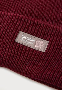 Maroon knitted beanie with a ribbed texture, featuring a rectangular logo patch in light gray with white text and a floral design.
