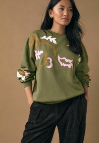 Pull vert olive avec broderie abstraite multicolore en rose, blanc et marron ; présente un col rond et des poignets côtelés. Pantalon en velours côtelé noir.