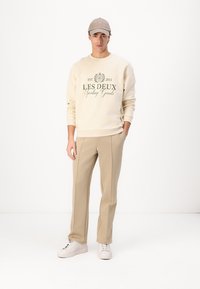 Beige sweatshirt met een donkergroene opdruk, gecombineerd met beige broeken. Witte sneakers maken de outfit af. Het model draagt een lichtgrijze pet.