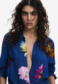 Floral print button-up shirt in denim blue, featuring large multicolored flowers, a relaxed fit, and long sleeves with a slight sheen.