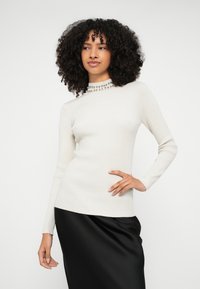 Λευκό πλεκτό πουλόβερ με ριγέ ύφασμα, μακριά μανίκια και ψηλό λαιμό διακοσμημένο με μεταλλικά στολίδια, σε συνδυασμό με μαύρη σατέν φούστα.