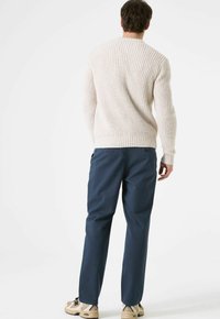 Mann trägt einen beigen gestrickten Pullover, dunkelblaue Hose und beige Sneaker, steht mit dem Rücken zur Kamera vor weißem Hintergrund.