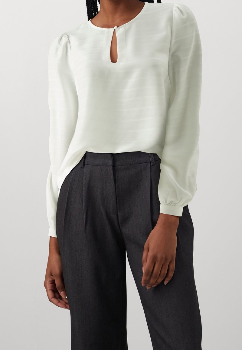 Blusa verde chiaro con un'apertura a chiave al collo, righe testurizzate e maniche a palloncino, abbinata a pantaloni grigi scuri su misura.