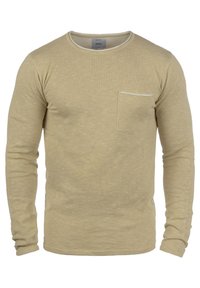 Beige pitkähihaista neulepusero, jossa on pyöreä pääntie ja pieni rintatasku vasemmalla puolella.