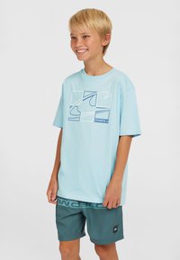 Ljusblå t-shirt med kort ärm och en geometrisk vit och blå grafisk design, kombinerad med turkosa shorts med en märkesresår.