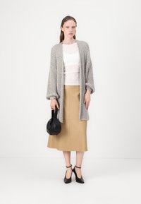 Sivý pletený blejzer s nadrozmernými rukávmi, biely čipkový top, béžová midi sukňa a čierna kabelka. Čierne sandále na podpätku dotvárajú tento outfit.