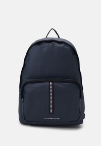 Zaino blu navy realizzato in tessuto liscio con una tasca anteriore con zip e una striscia decorativa. Ha un manico in cima e un logo nella parte inferiore.