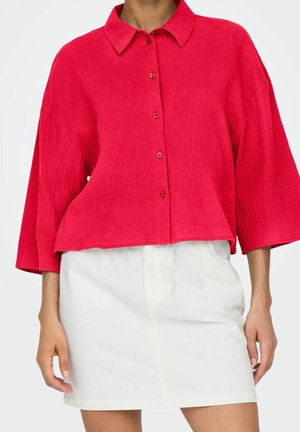 Mujer con camisa roja holgada abotonada, mangas anchas y falda blanca de cintura alta, de pie con los brazos relajados a los lados.