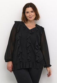 Sort bluse med V-hals, med flæsedetaljer langs fronten og ærmerne, samt en flæse i kanten. Letvægtigt, semi-transparent stof.