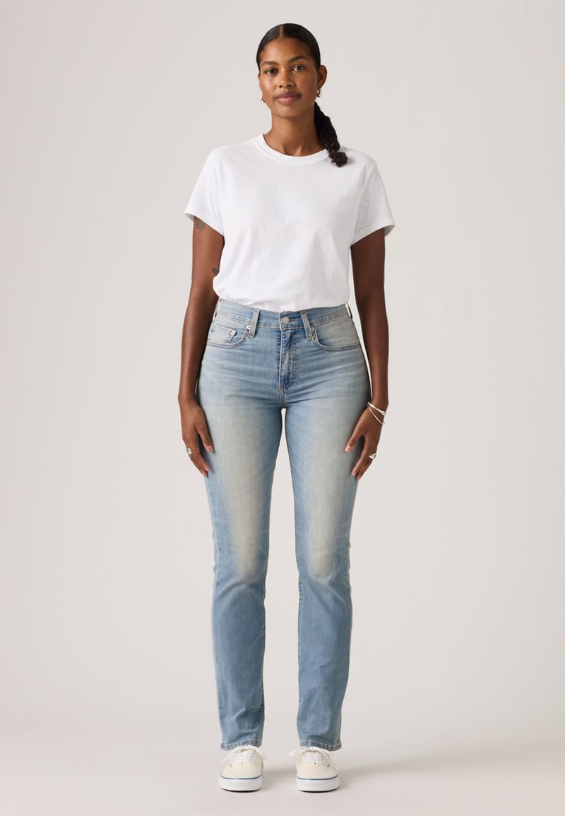 Femme debout, faisant face à l'avant, portant un t-shirt blanc, un jean taille haute bleu clair, des baskets blanches et des bijoux argentés, sur un fond uni.