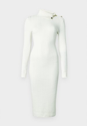 Fehér bordázott kötött ruha magas gallérral, aszimmetrikus nyakkivágással és díszes aranyszínű fém kiegészítőkkel a vállán. Testhez simuló sziluett.