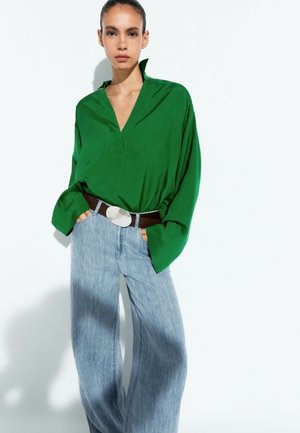 Frau mit zurückgebundenem Haar trägt eine lockere grüne Bluse und weite hellblaue Jeans mit braunem Gürtel und großer ovaler Schnalle, Hände in den Taschen.