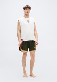 Λευκό αμάνικο μπλουζάκι με μικρό γραφικό, συνδυασμένο με χακί πράσινα σορτς με μπεζ διακοσμητικές λεπτομέρειες και διακριτικό λογότυπο στο πλάι.