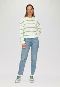 Weiße Strickjacke mit grünen Streifen, Rundhals-Ausschnitt und weiten Ärmeln, kombiniert mit hellblauen Jeans und Sneakers mit grünen Akzenten.